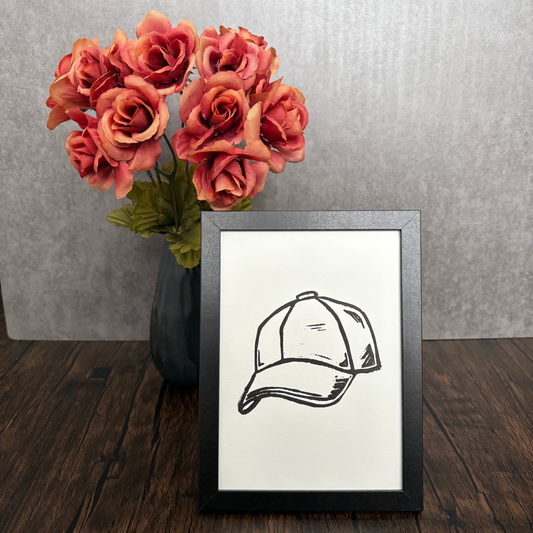Framed black and white drawing of a cap next to a vase of pink roses on a wooden surface.