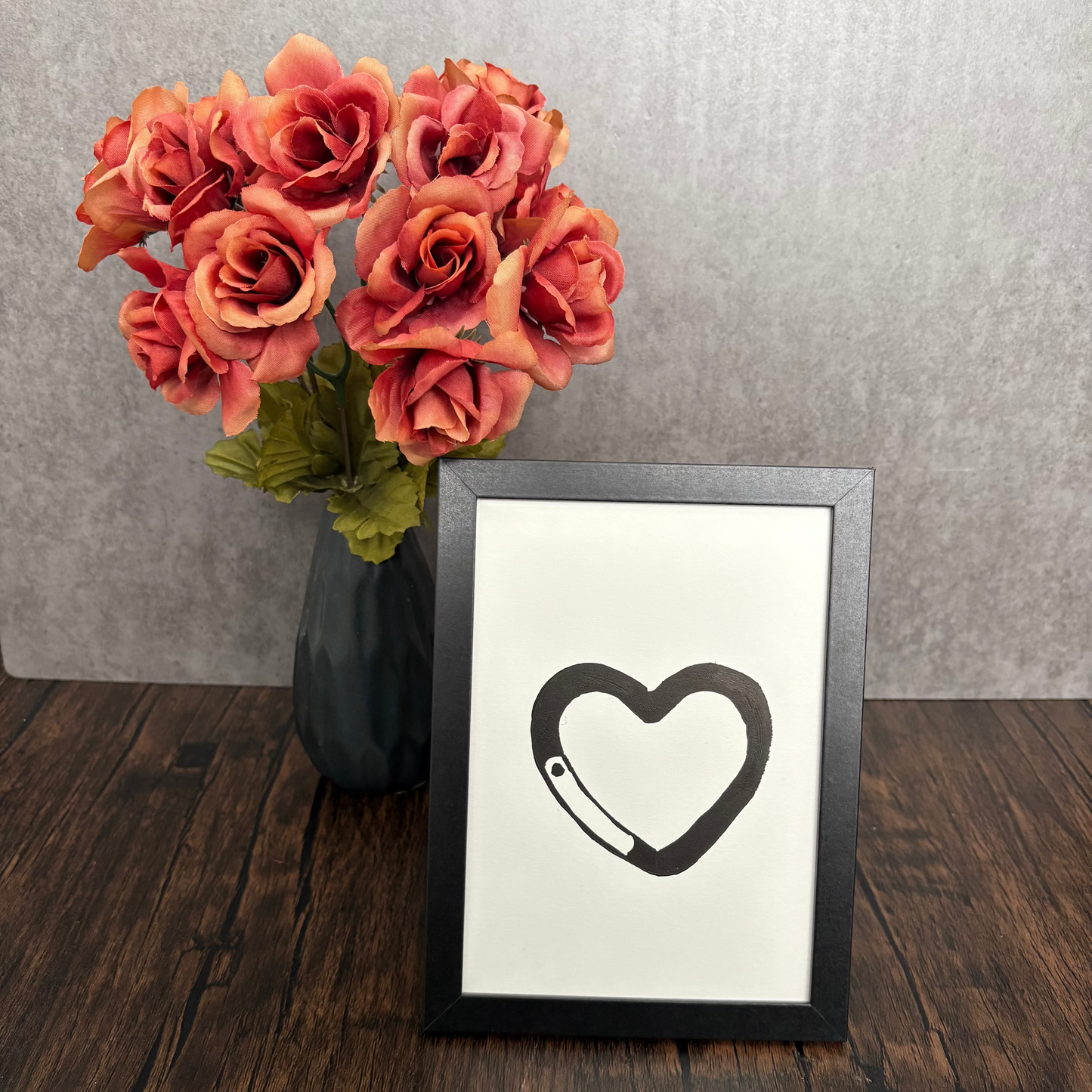 Framed artwork with a heart design next to a vase of pink roses on a wooden surface.