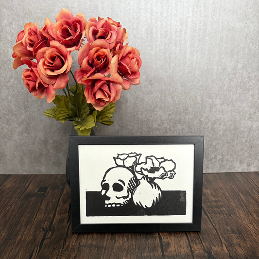 Framed artwork of a skull with roses next to a vase of red flowers on a wooden surface.