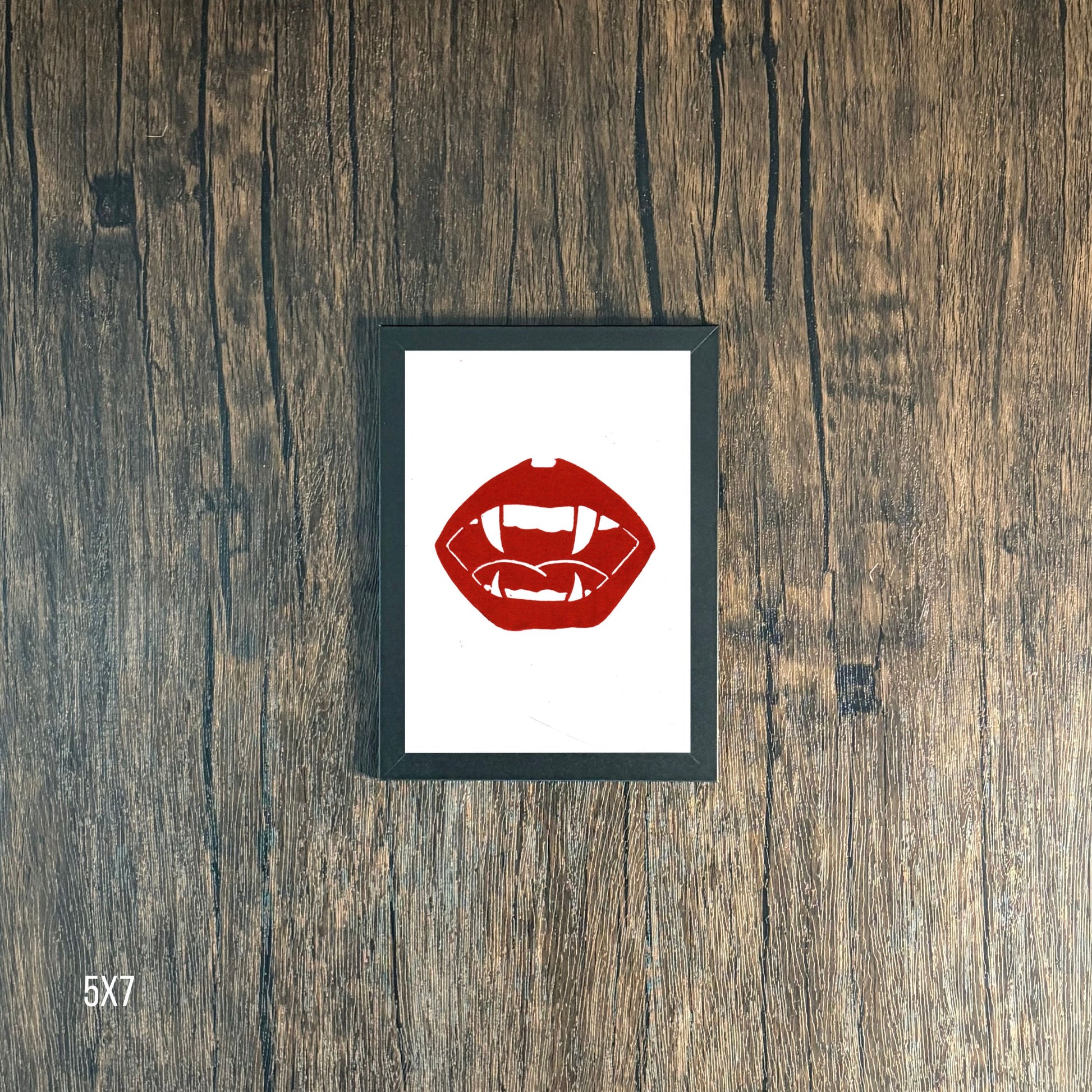 Framed Red lips and vampire fangs graphic on white  paper against a wooden background