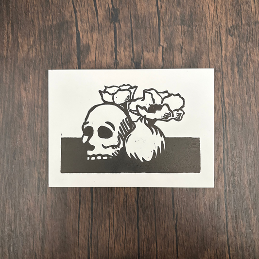 Linocut print of a skull, flowers, and pomegranate on a dark background.