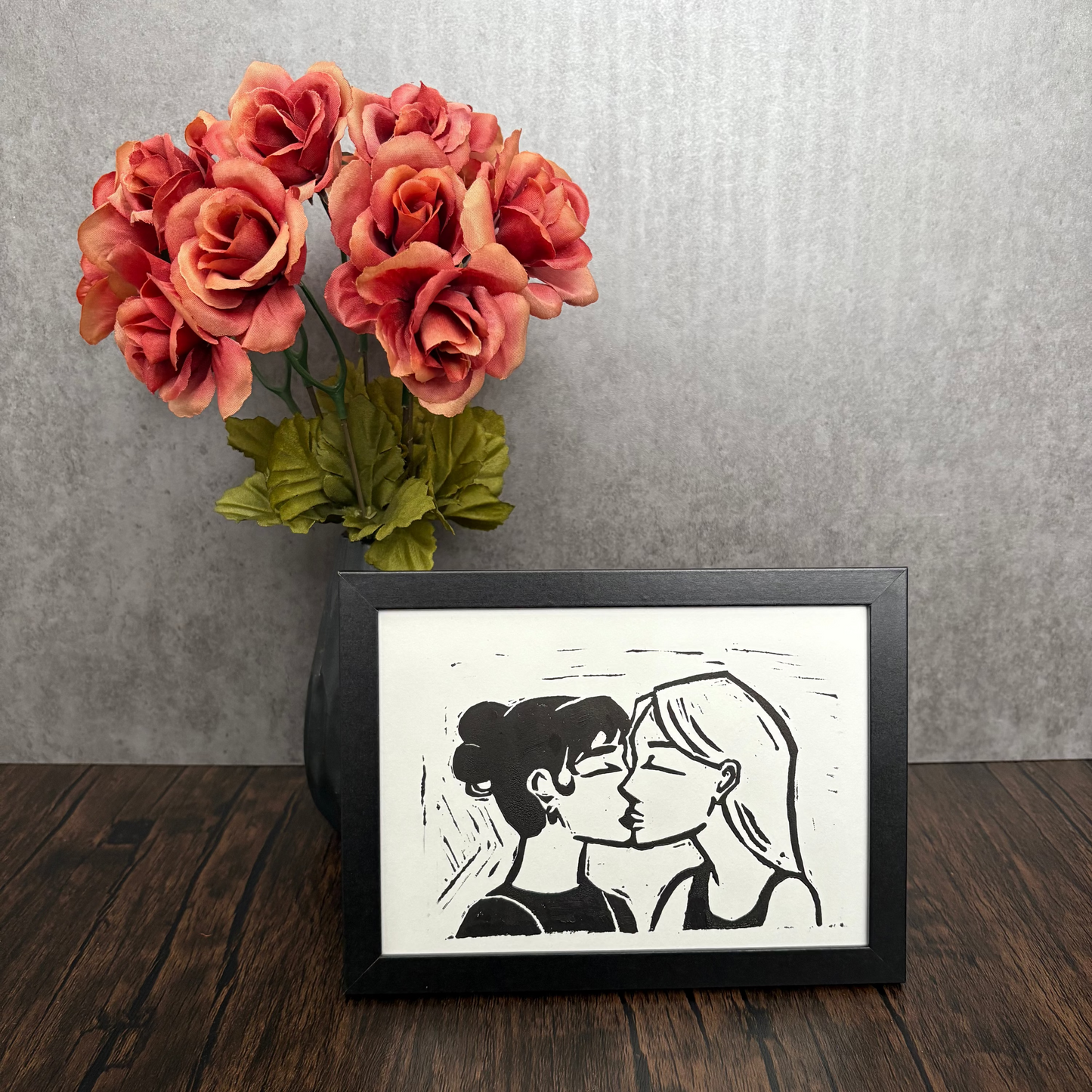 Framed artwork of two faces on a wooden surface with pink flowers in the background